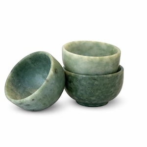 May include: Three small, hand-carved jade bowls in varying shades of green. One bowl is tilted on its side, while the other two are stacked. The bowls have a smooth, polished finish and a natural, organic appearance.