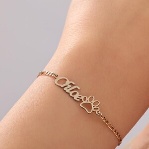 Pode incluir: Uma pulseira dourada com o nome "Chloe" em escrita cursiva e um berloque de pegada de pata. A pulseira está em um pulso, em um fundo neutro.