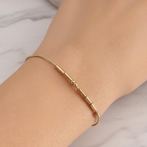 Peut inclure: Un bracelet en or délicat avec une série de petites perles cylindriques et une fine chaîne. Le bracelet est présenté sur un poignet, mettant en valeur son design minimaliste et son élégance subtile. La longueur du bracelet est d'environ 18 cm.