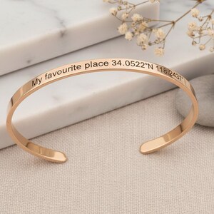 May include: A rose gold-toned metal cuff bracelet with a smooth, polished finish. The outer surface is engraved with the black text "My favourite place 34.0522°N 118.2431°". The bracelet features an open-ended design with rounded, flat ends.