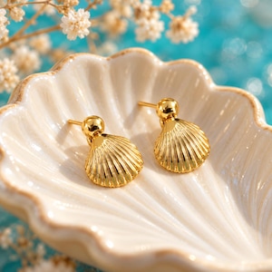 May include: A pair of gold-toned stud earrings shaped like small seashells with vertical ridges. The earrings feature a polished metallic finish and are resting inside a white ceramic dish molded into the shape of a large scallop shell.