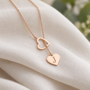 May include: A rose gold-toned necklace featuring a delicate chain that passes through an open heart-shaped charm, ending in a solid heart-shaped pendant engraved with the capital letter J in a classic serif font style.