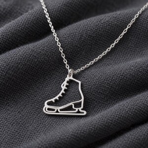 Puede incluir: Un collar de tono plateado con un colgante calado en forma de patín de hielo. El colgante cuelga de una cadena fina de tono plateado a juego, mostrando el contorno limpio de la bota de patinaje y la cuchilla metálica.