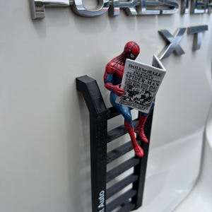 May include: A Spider-Man figurine in red, blue, and black, reading a newspaper, sitting on a black ladder. The newspaper reads "Daily Bugle" and "Friendly Neighborhood Spider-Man". The ladder is attached to a white car.