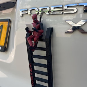 Può includere: Una statuetta di Deadpool rossa e nera seduta su una scala nera attaccata a un veicolo. La statuetta tiene in mano un tablet bianco. La scala ha il testo "FIGURE IT AUTO" in rosso.