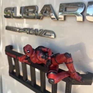 Può includere: Un action figure di Deadpool rosso e nero in posa reclinata, montato su un accessorio in plastica nera a forma di scala. La figura è posizionata contro una superficie bianca del veicolo con emblemi cromati Subaru e AWD con testo.