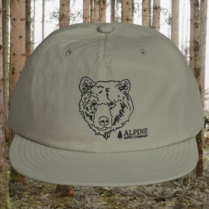 May include: A light green baseball cap with a black bear graphic and the text "ALPINE SUPPLY COMPANY". The cap has a curved brim and a button on top. The background is a forest.