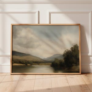 May include: A framed landscape painting featuring a serene river scene with mountains in the background. The artwork has a muted color palette of greens, browns, and blues, with a wooden frame. The painting evokes a sense of tranquility.