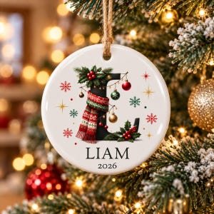 Puede incluir: Un adorno navideño redondo de cerámica blanca colgado de un cordel de yute. El centro presenta una letra L negra grande decorada con una bufanda de punto roja y verde, acebo y pequeñas bolas. El nombre LIAM y 2026 están impresos debajo.