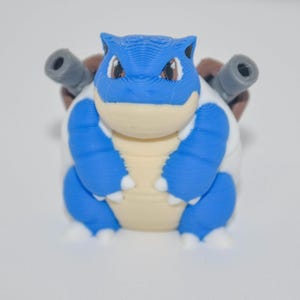 May include: A 3D printed figurine of a blue creature with a cream-colored belly, white accents on the hands and feet, and two gray cannons protruding from a brown shell on its back. The figure features visible horizontal layer lines from the printing process.