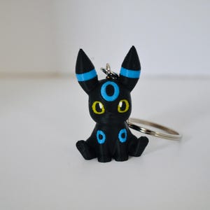May include: A black keychain figure of a creature with pointed ears, featuring bright blue circular markings on its forehead, ears, and legs, along with large yellow eyes. The figure is attached to a silver metal split ring for keys.