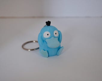 Op de afbeelding: Een lichtblauwe sleutelhanger van een klein, eendachtig wezen met een brede snavel, grote witte ogen met zwarte pupillen en drie kleine zwarte plukjes op het hoofd. Het figuurtje zit in een zittende houding met de handen op het hoofd.