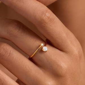 May include: A gold ring featuring a small, round, white gemstone. The ring has a minimalist design, with a slender band that curves around the finger. The gemstone is set in a delicate prong setting.