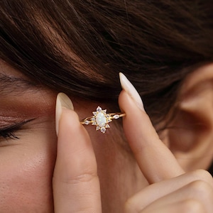 Peut inclure: Une bague en or avec une pierre centrale ovale blanc cassé, entourée de petites pierres transparentes. La bague présente un design délicat en forme d'éclat d'étoile. La bague est tenue par les doigts d'une personne.