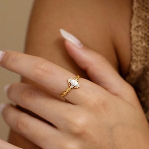 May include: A gold ring with a white gemstone in a marquise shape. The ring has a delicate, textured band. The ring is on a person's finger, with a neutral background.