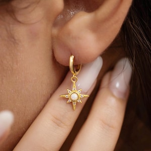 Könnte beinhalten: Ein goldfarbener Ohrring mit Sternenmuster, mit einem zentralen weißen Stein und kleinen weißen Akzenten. Der Ohrring ist an einem kleinen Reifen befestigt. Das Bild zeigt den Ohrring an einem Ohr.