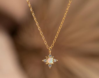 Pode incluir: Um delicado colar de ouro com um pingente em forma de estrela, com uma pedra de opala central e cristais transparentes ao redor. O pingente pende de uma fina corrente de ouro, criando um design simples, mas elegante.