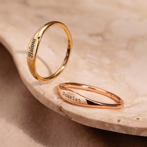 Peut inclure: Deux bagues personnalisées, une en or et une en or rose, chacune gravée d'un nom. La bague en or porte l'inscription "Briana", tandis que la bague en or rose porte l'inscription "Scarlet". Les bagues sont présentées sur une surface claire et texturée.