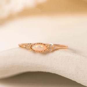 May include: A delicate gold ring featuring an oval opal center stone. The ring has small, sparkling accents on either side of the opal. The ring is set on a white surface, with a soft, blurred background.
