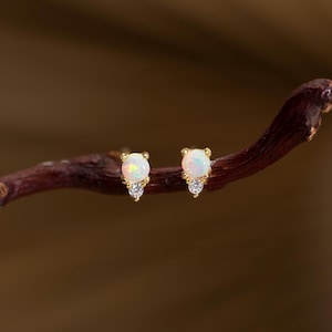 Peut inclure: Une paire de boucles d'oreilles à clous en or avec une pierre d'opale blanche ronde et un petit accent en cristal transparent. Les boucles d'oreilles sont présentées sur une branche brune foncée. Bijoux.