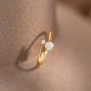 May include: A delicate gold ring featuring two white opal gemstones. The ring has a minimalist design with a small opal and a larger, round opal. The ring is set against a soft, neutral background.