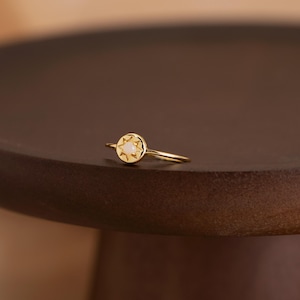 Peut inclure: Une bague en or avec une face circulaire ornée d'un motif en forme d'étoile et une petite pierre claire au centre. La bague est posée sur une surface en bois brun foncé, créant un contraste de couleurs et de textures. Le design délicat de la bague suggère un bijou.