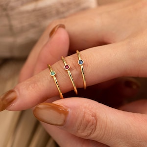 Puede incluir: Tres delicados anillos de oro, cada uno con una pequeña gema de color: verde, rojo y azul. Los anillos se muestran en un dedo, mostrando su diseño minimalista y acentos vibrantes. Una pieza de joyería elegante.