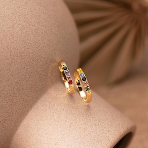 May include: Two gold hoop earrings, each featuring a line of colourful gemstones. The gemstones include purple, red, yellow, blue, and green. The earrings are presented on a neutral surface, with a blurred background.
