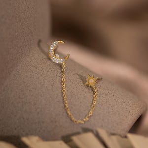 Op de afbeelding: Een gouden oorbel met een maanvormig ontwerp, bezet met kleine, heldere stenen. Een fijne ketting verbindt de maan met een stervormige oorbel met een kleine, ronde, crèmekleurige steen. De oorbel wordt gepresenteerd tegen een neutrale achtergrond.