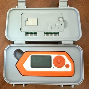May include: A Flipper Zero portable multi-tool device with an orange and white casing, featuring a screen and circular navigation buttons, housed inside a grey protective carrying case with molded compartments for accessories and components.