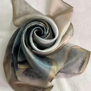 May include: A silk scarf with an abstract design in shades of blue, grey, and beige. The scarf is folded and arranged to create a layered, textured effect, with a soft, lustrous sheen. The edges are finished with a subtle border.