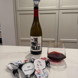 May include: A bottle of The Pinot Project wine with a wine stopper that says "The Hamptons". Several decorative wine stoppers with various designs and a glass of red wine are on a white surface.