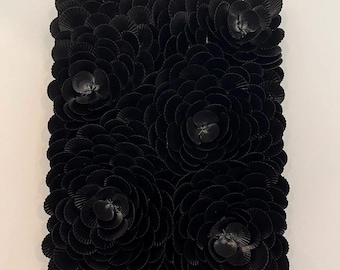 Könnte beinhalten: Ein quadratisches, schwarzes Wandkunstwerk mit einer strukturierten Oberfläche, die aus zahlreichen kleinen, fächerförmigen Elementen besteht. Fünf große, stilisierte Blumenformen sind über die Oberfläche angeordnet, jede mit einem detaillierten Zentrum.