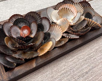Può includere: Un vassoio decorativo in legno con una composizione floreale realizzata con conchiglie. Le conchiglie sono in tonalità di marrone, beige e bianco, con una piccola conchiglia arancione vibrante al centro. Il vassoio ha una cornice rettangolare marrone scuro.