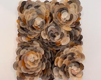 Può includere: Un'opera d'arte murale quadrata composta da numerose conchiglie disposte a formare quattro grandi fiori. Le conchiglie presentano una gamma di colori neutri, tra cui beige, marrone chiaro e grigio, con alcuni accenni di arancione. L'opera è montata su uno sfondo bianco.
