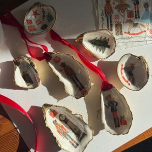 May include: Hand-painted oyster shell Christmas ornaments with gold trim and red ribbon. Each shell features a different festive design, including a Christmas tree, gifts, and nutcrackers. These unique ornaments add a touch of coastal charm to holiday decor.