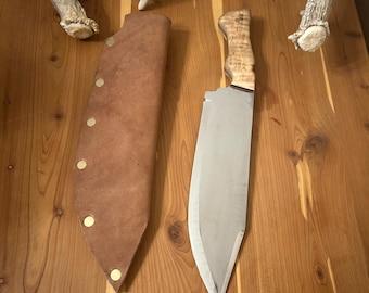 May include: A large, stainless steel cleaver with a light-colored wooden handle and a brown leather sheath. The sheath is secured with brass-colored rivets. The knife is displayed on a wooden surface, with a pair of antlers in the background.
