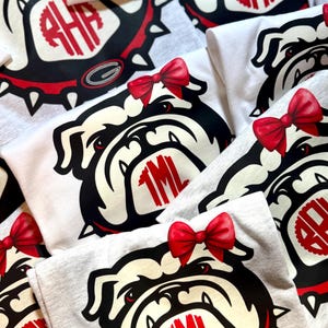 May include: Several white and grey t-shirts with a bulldog graphic and a red bow. The bulldog design includes a red monogram. These shirts are likely for a sports team or school.