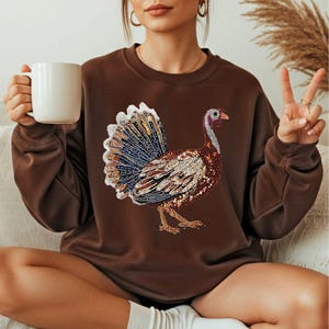 May include: A brown sweatshirt featuring a detailed, colorful turkey design. The turkey is embellished with various colors and textures, including blue, brown, and gold. The sweatshirt is a solid brown color and has long sleeves.