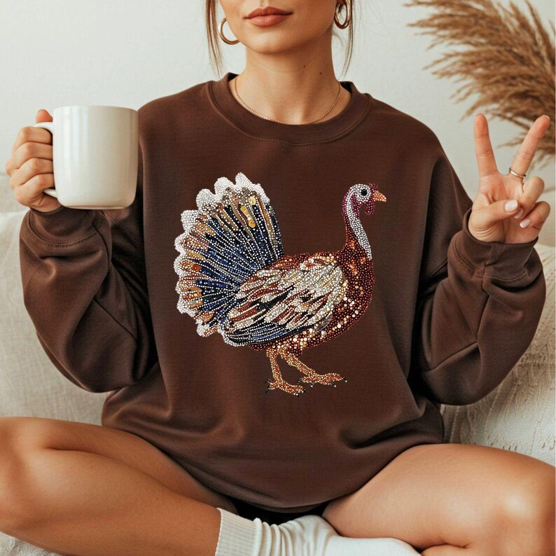 May include: A brown sweatshirt featuring a detailed, colorful turkey design. The turkey is embellished with various colors and textures, including blue, brown, and gold. The sweatshirt is a solid brown color and has long sleeves.