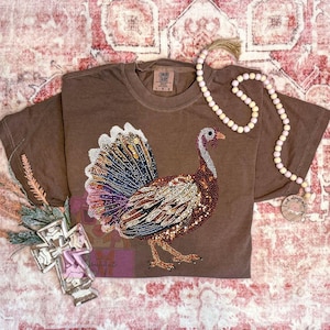 May include: Brown short-sleeved t-shirt with a large, embellished turkey design. The turkey features a colorful, jeweled tail and body. The shirt has a Comfort Colors tag and is labeled L.G.G.