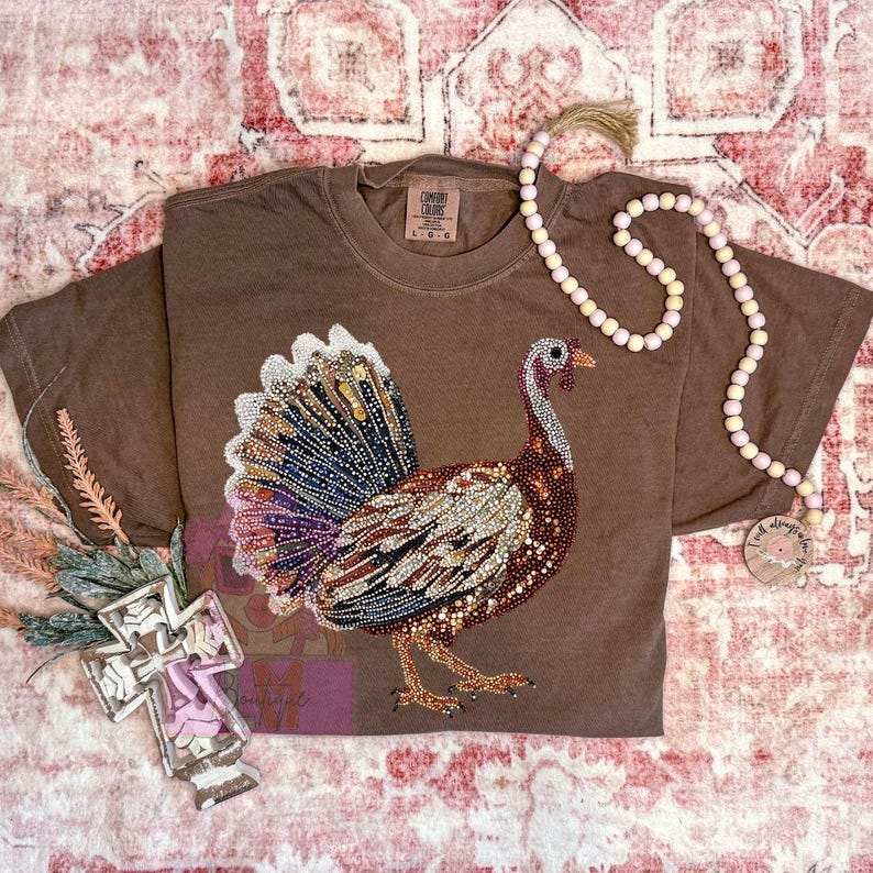 May include: Brown short-sleeved t-shirt with a large, embellished turkey design. The turkey features a colorful, jeweled tail and body. The shirt has a Comfort Colors tag and is labeled L.G.G.