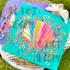 May include: Two colorful t-shirts with seashell designs. One is teal, the other is purple. The teal shirt features a large, multi-colored seashell with pink, yellow, and blue hues, surrounded by colorful confetti. The purple shirt has a similar design.