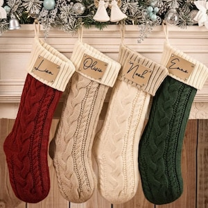 May include: Four cable-knit Christmas stockings in red, beige, cream, and green hang from a mantelpiece. Each stocking has a cream-coloured cuff and a small leather tag with a name: Liam, Olivia, Noah, and Emma. Festive holiday decor.