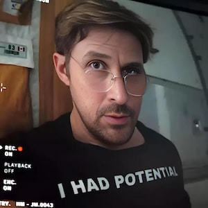 Könnte beinhalten: Eine Person trägt ein schwarzes T-Shirt mit dem weißen Text "I HAD POTENTIAL". Die Person trägt goldrandige Brille. Der Hintergrund umfasst einen Bildschirm und einige Geräte.