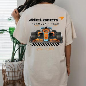 Könnte beinhalten: Cremefarbenes T-Shirt mit einem McLaren Formula 1 Team-Design. Die Grafik zeigt das McLaren-Logo, ein orange-blaues Formel-1-Auto und den Text "Papaya and Black".