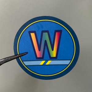 May include: A round sticker with a blue background and a large, colorful "W" in the center. The "W" transitions from red to yellow and green. A yellow and blue border surrounds the design.