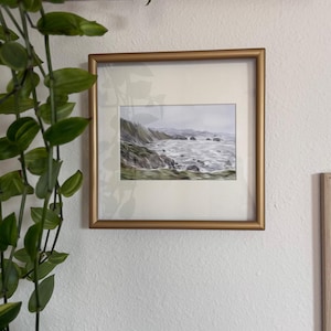 May include: A framed landscape painting of a coastal scene with a gold frame. The artwork depicts a rocky shoreline with crashing waves and a cloudy sky. Green foliage is visible on the cliffs. The painting is mounted on a white wall.
