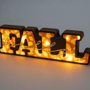 May include: A brown, illuminated sign spelling "FALL". The letters are outlined in brown and filled with a warm, glowing light. The interior of the letters features a leaf pattern, and a white cursive script is draped across the letters.