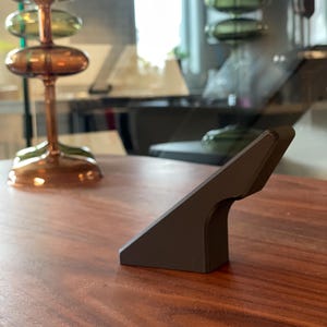 Puede incluir: Una manija de puerta triangular negra con un borde redondeado. La manija está hecha de un material liso y mate y está montada en una superficie de madera.